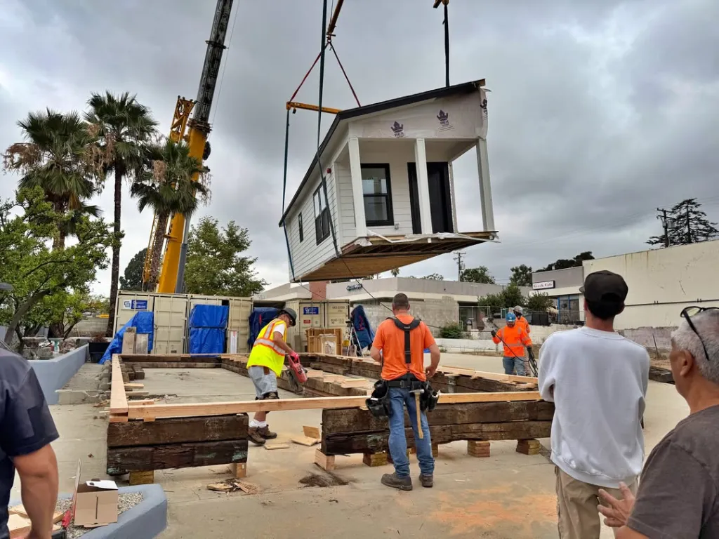 A crane lifting a model home onto a wooden foundation with workers observing.