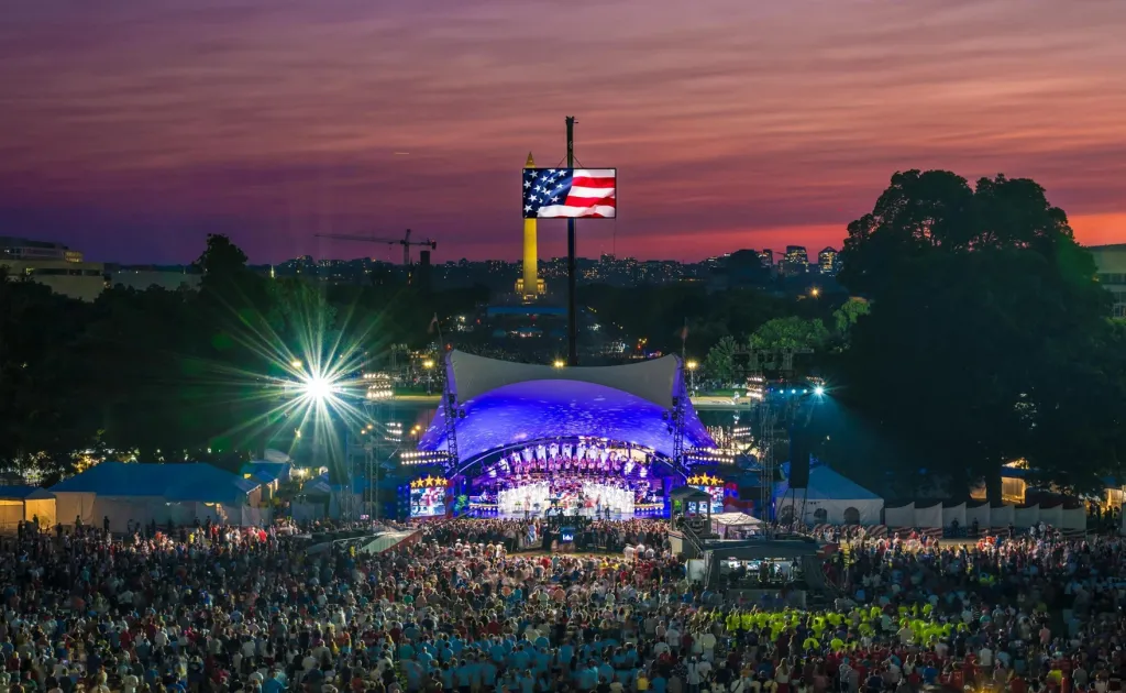 A large crowd gathered for A Capitol Fourth concert, with a bandshell and an American flag screen, against a twilight sky with the Washington Monument in the distance.