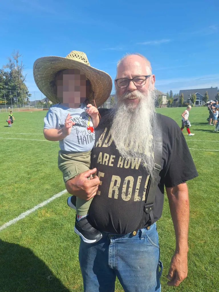 Carl McDaniel, a man with a long white beard and glasses, holding a baby in a straw hat on a grassy field.