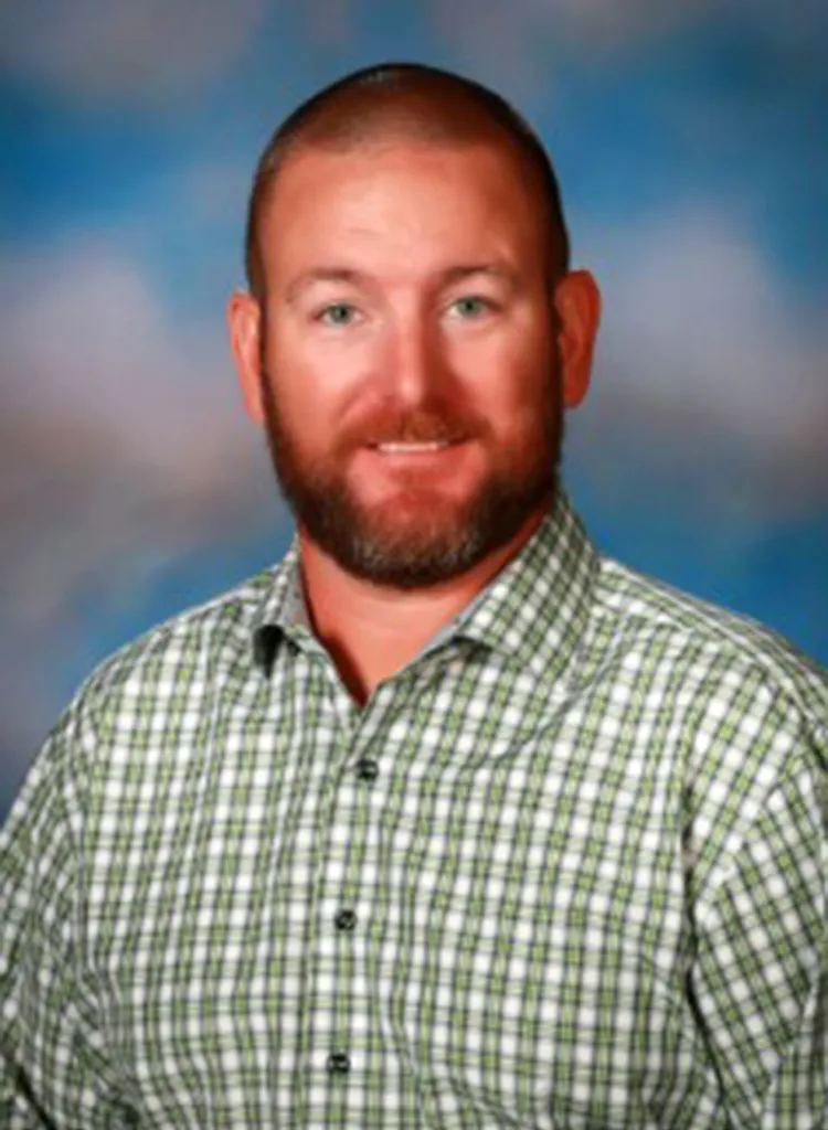 Richard Aspinwall, a man with a reddish-brown beard and a shaved head, smiles in a school photo, wearing a green and white plaid shirt.