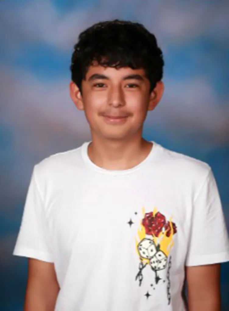 Christian Angulo, a young man with dark, curly hair, smiles slightly while wearing a white t-shirt with a flaming rose and dice graphic.