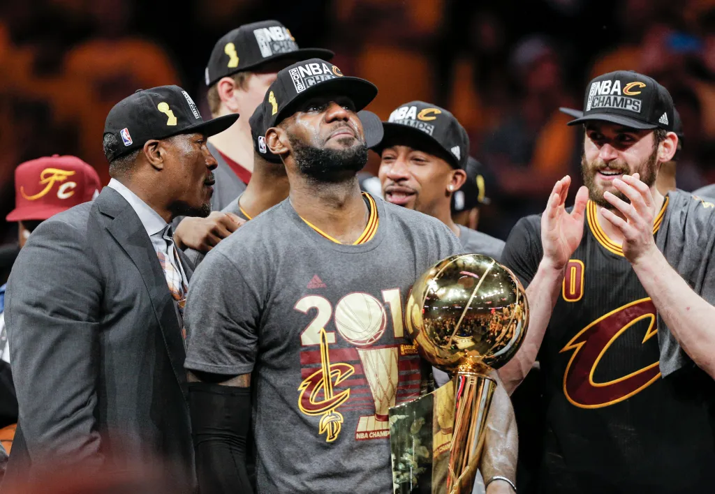 LeBron James holding the Larry O’Brien NBA Championship Trophy while wearing an NBA Champions hat and a 