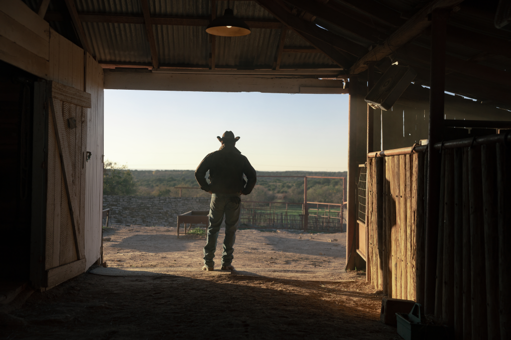 Cole Hauser as Rip Wheeler, seen from behind, stands in a barn looking out at the Dutton Ranch.