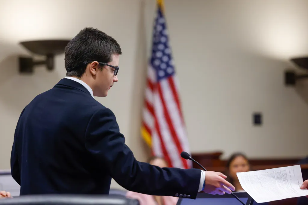 Colt Gray, a 16-year-old high school shooting suspect, extends his hand to receive a document in court with an American flag in the background.