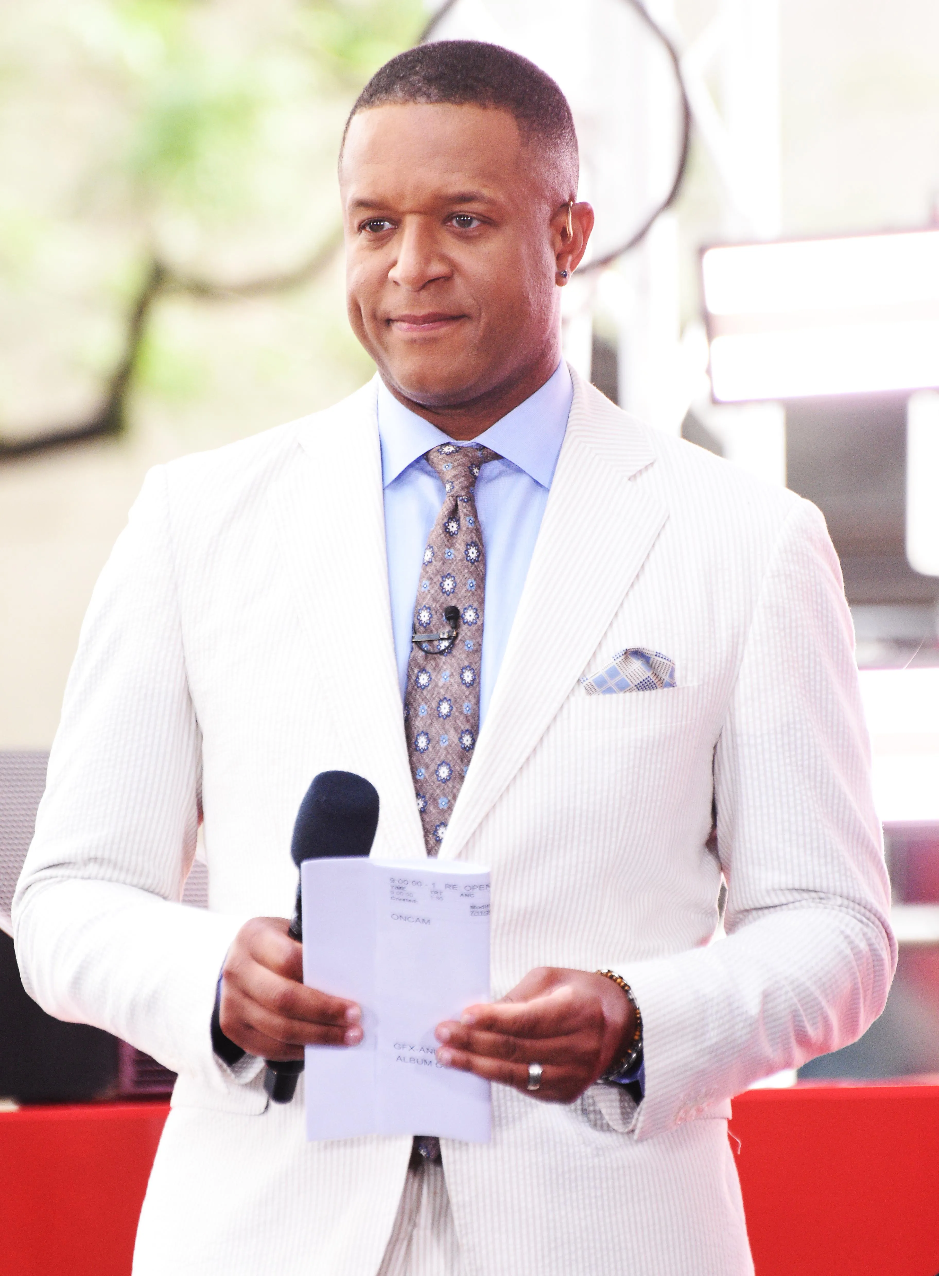 Craig Melvin wearing a white suit, light blue shirt, and patterned tie, holding a microphone and papers with 