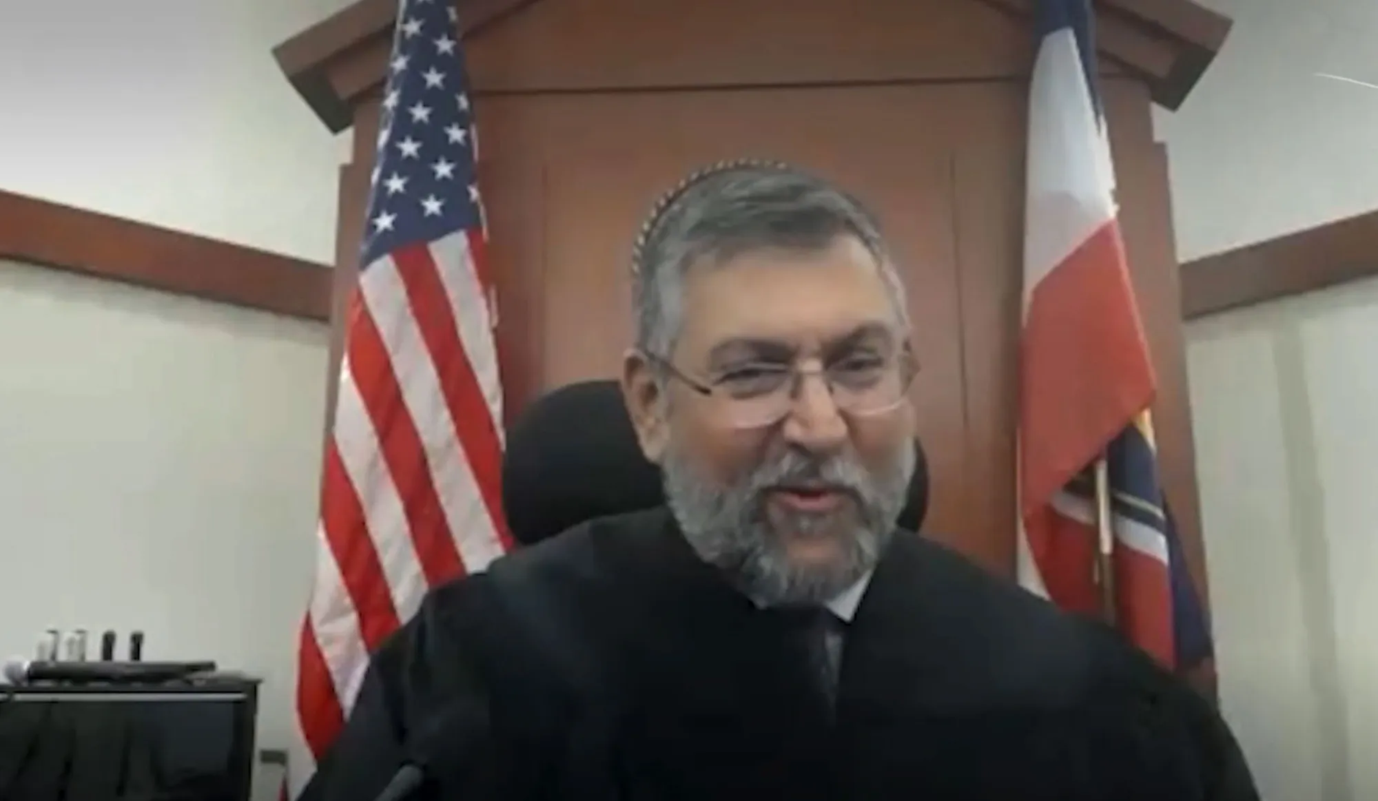 Dakota Mortensen, an older man with a beard and glasses, smiles while wearing a black judicial robe, with American and Texas flags behind him.