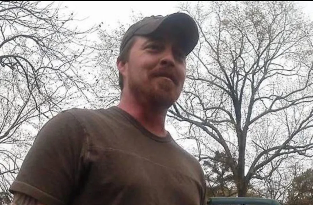 Daniel Robbins, a man with a beard and baseball cap, looks at the camera against a backdrop of bare trees.