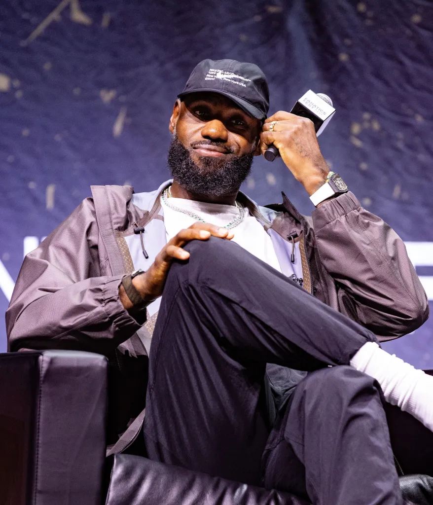 LeBron James smiling while sitting in a chair, wearing a black hat, a grey jacket, a white t-shirt, and black pants.