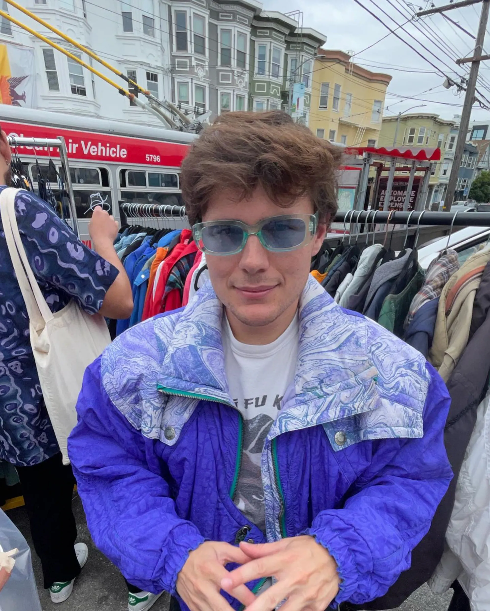 Robert Capron wearing a purple jacket and light-blue sunglasses, standing in front of a clothing rack and a trolley bus.