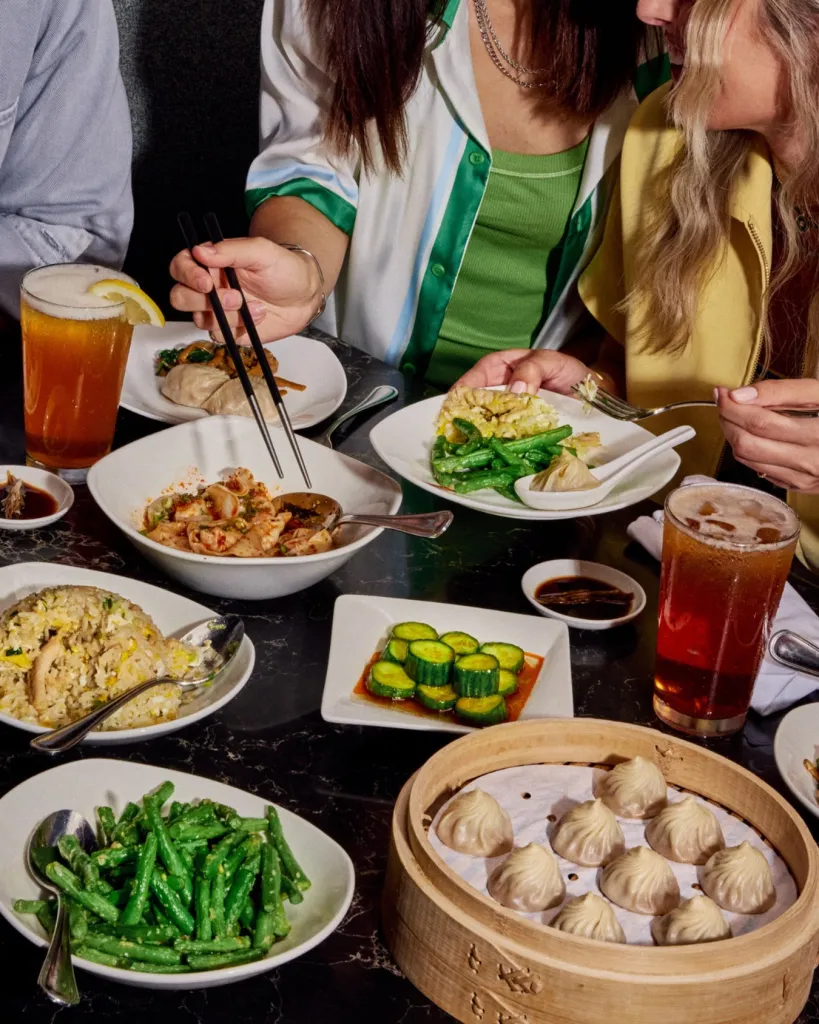 Diners enjoying a meal of various dishes including dumplings, green beans, cucumbers, and fried rice, with drinks.