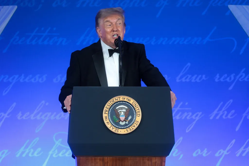 Donald Trump speaking at a podium with the Seal of the President on it, against a blue background with faint script.