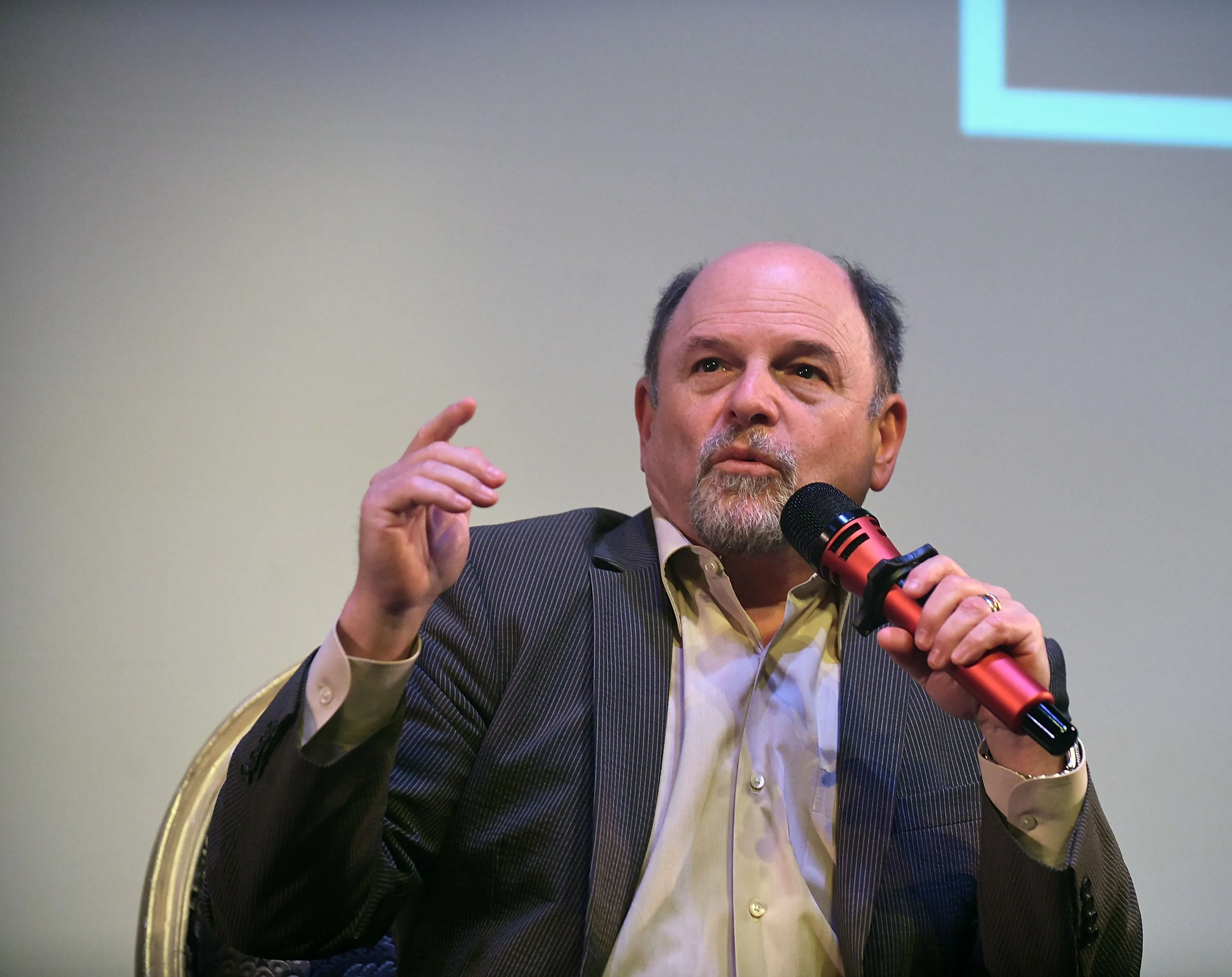 Jason Alexander speaking at an event, holding a red and black microphone in one hand and gesturing with the other.