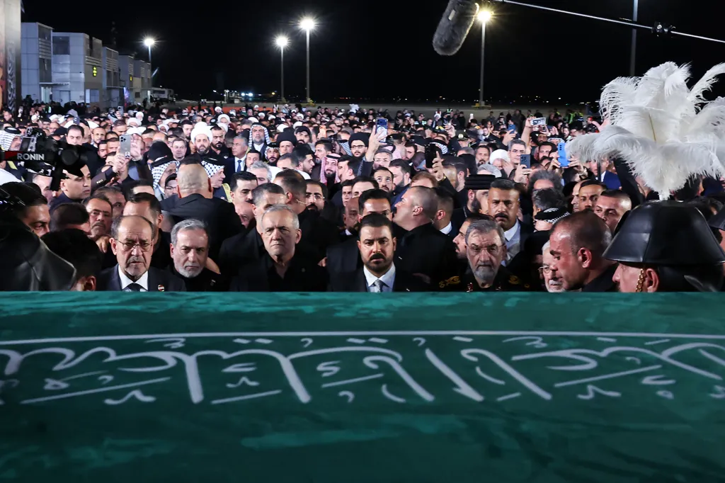 A crowd of men surrounds a green coffin at an airport at night, with two bright lights in the background.