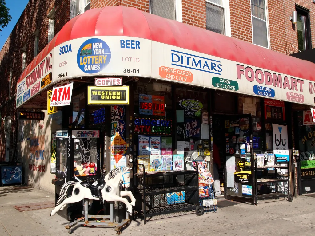 The exterior of Ditmars Foodmart in Queens, a convenience store with a red awning and signs for lottery, beer, sodas, and various other goods, and a kiddie ride of a black and white horse.