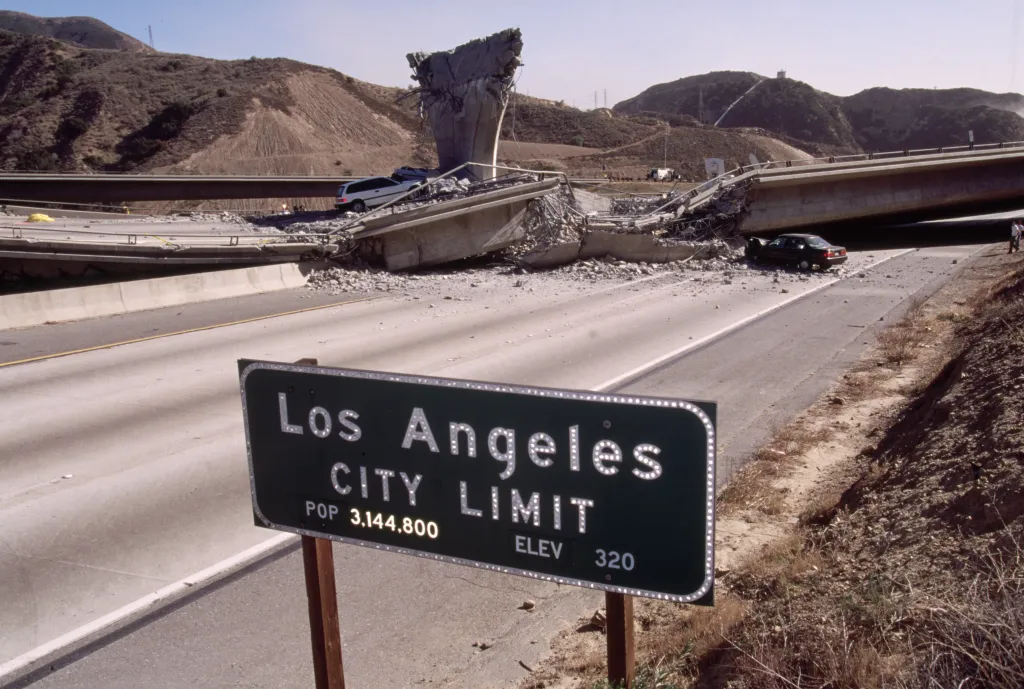 Road sign for Los Angeles in the foreground, with a freeway overpass destroyed by the Northridge earthquake in the background.
