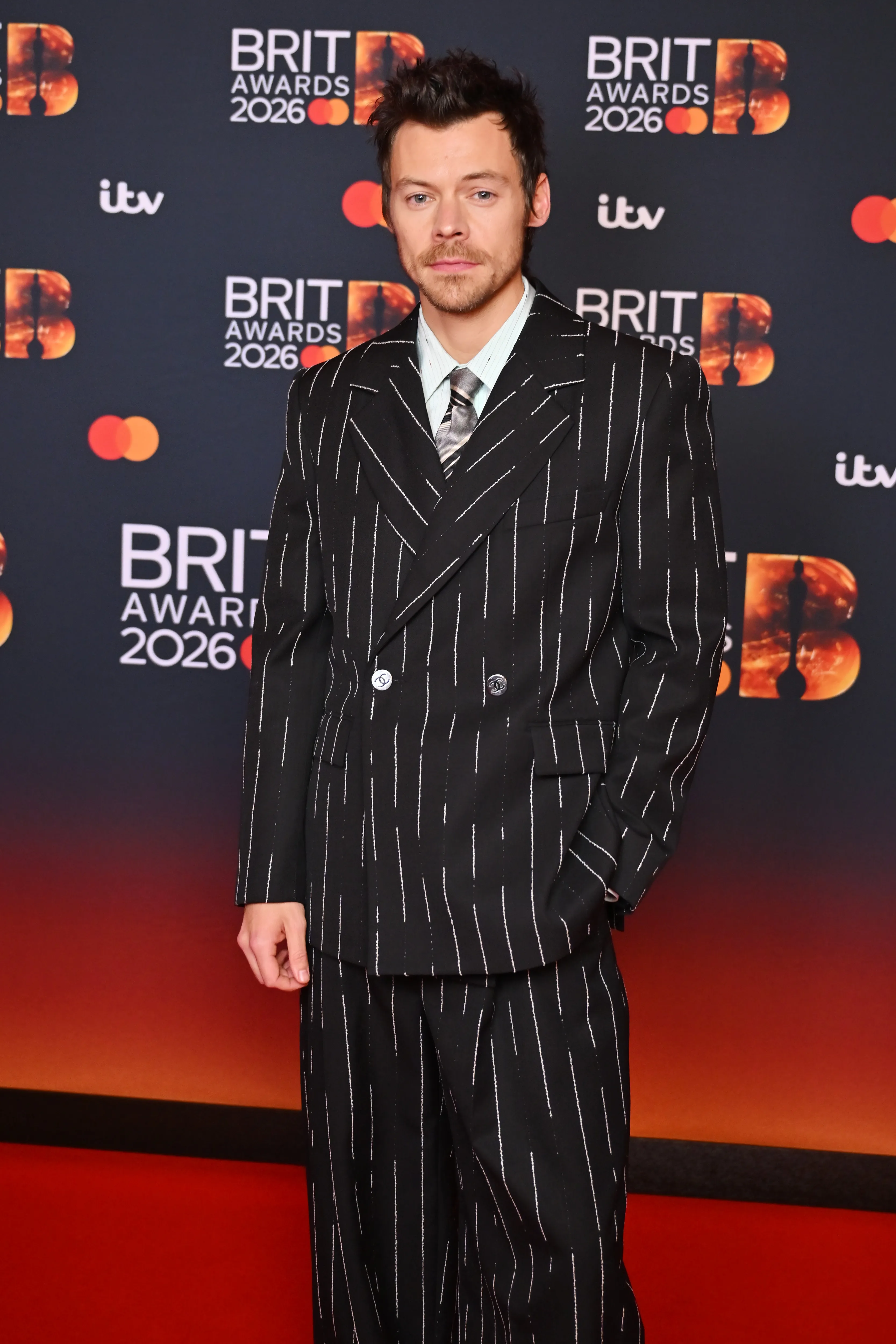 Harry Styles in a black pinstripe suit, light green shirt, and striped tie, standing against a backdrop of 