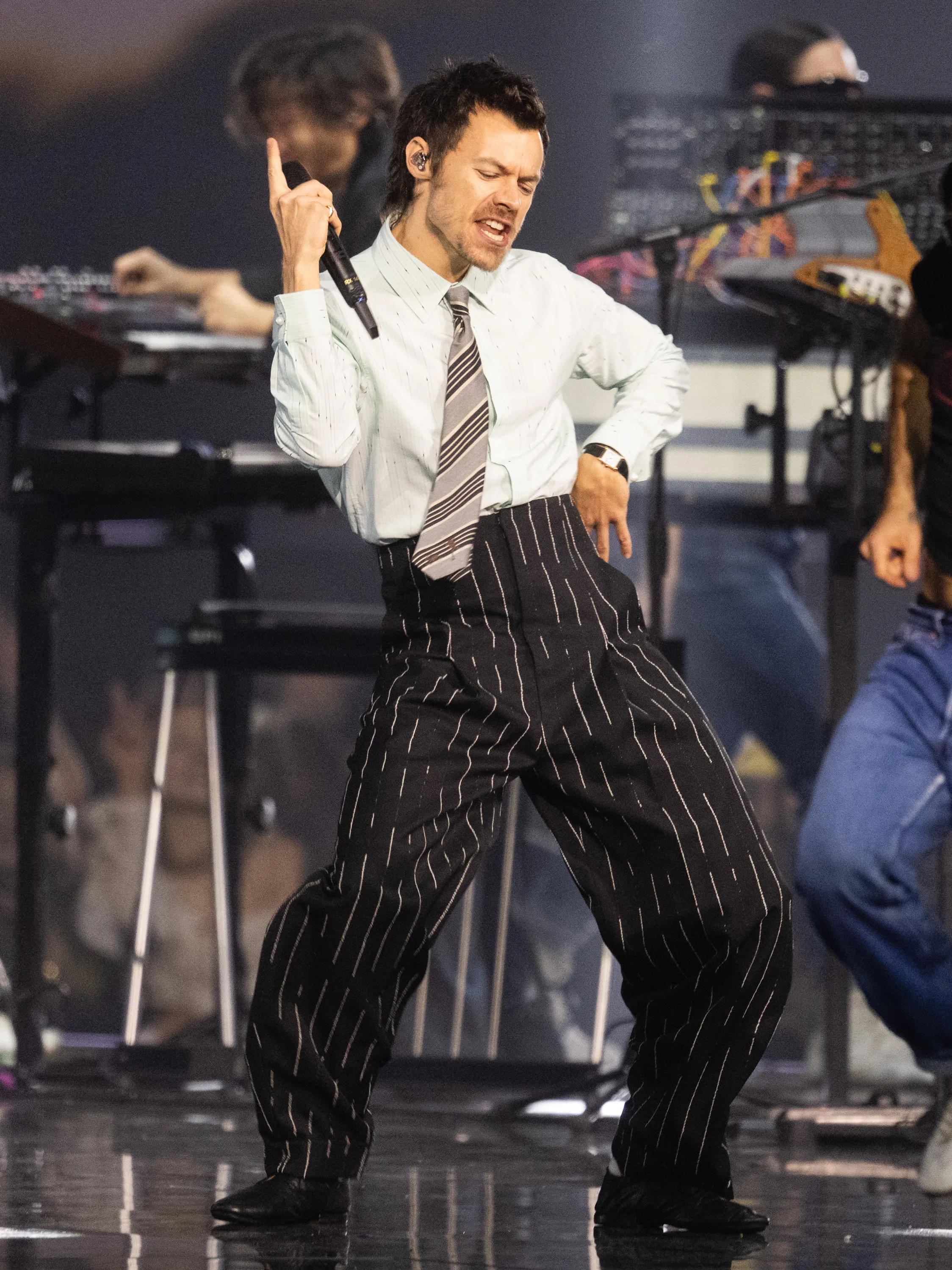 Harry Styles performs on stage with a microphone in hand, wearing a striped shirt, tie, and pinstripe pants.