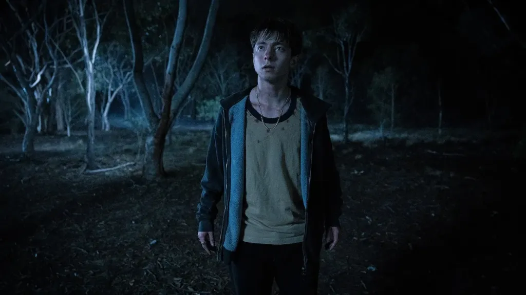 Joe Bird stands alone in a dark forest looking at something out of frame in a still from 