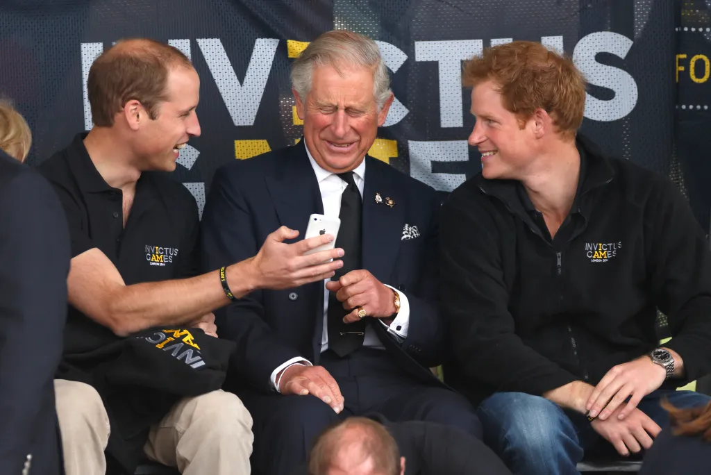 Prince William, in a black top with the Invictus Games logo showing a mobile phone to Prince Charles , in a navy blue suit, while Prince Harry, in a navy blue sweater with Invictus logo, watches.