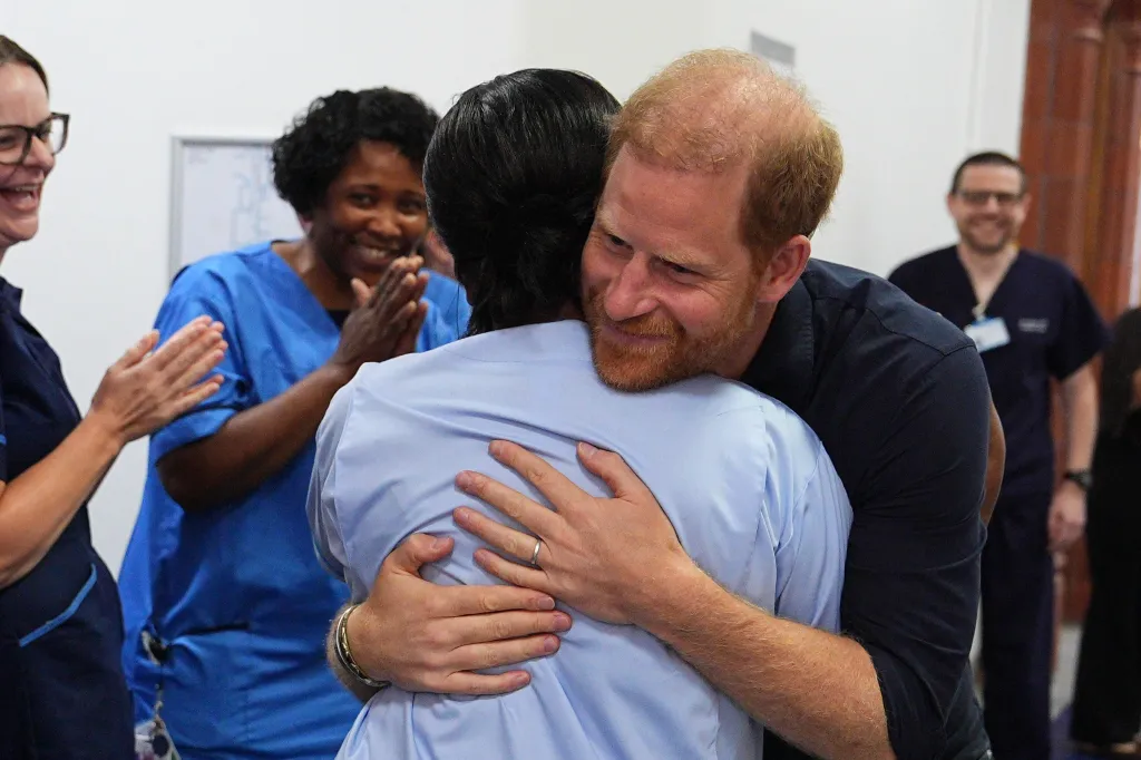 Prince Harry, in a blue shirt, hugging a nurse, in a blue shirt, with other medical staff smiling and clapping in the background.