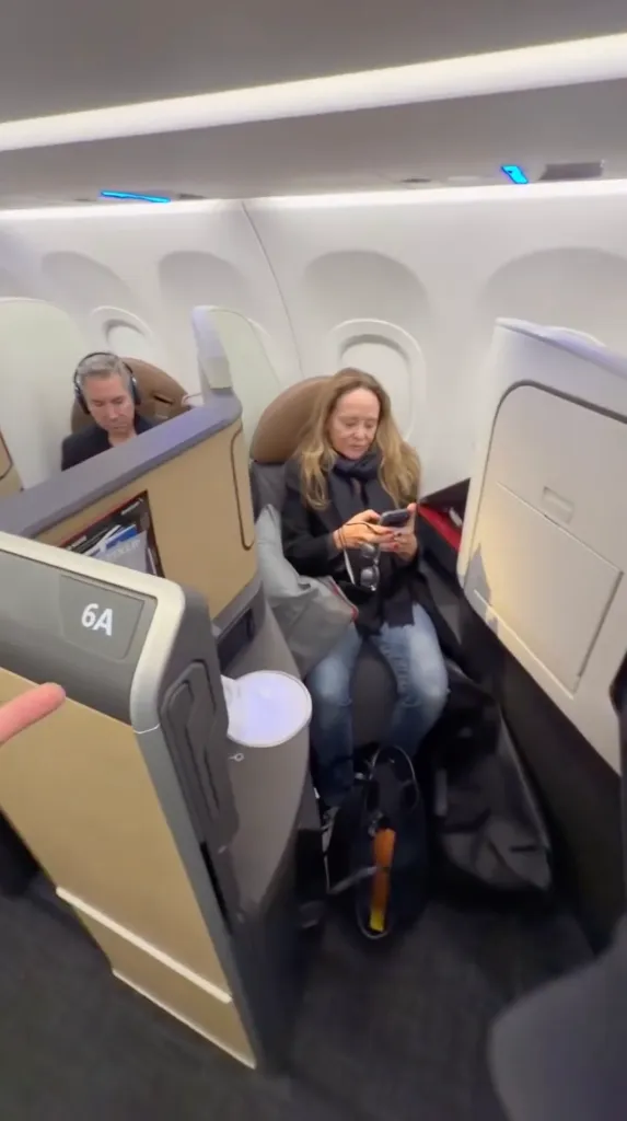 A woman sitting in a first-class airplane seat using her phone, with a man in the seat behind her.