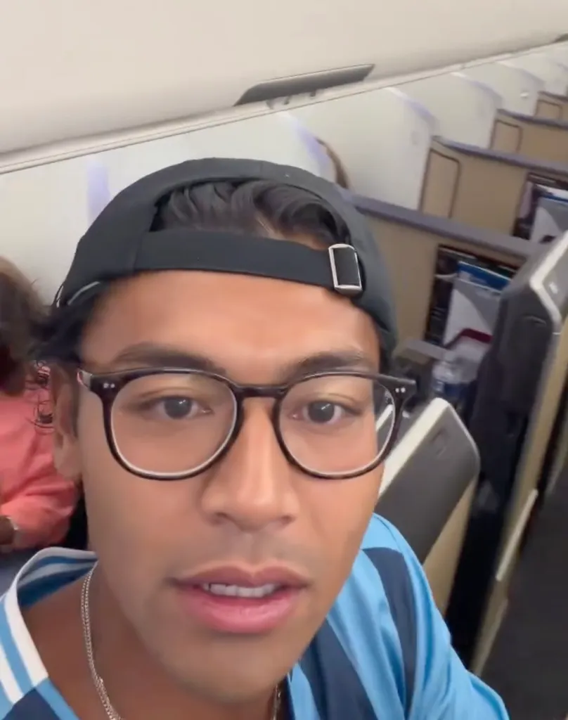 A man wearing glasses and a cap, filming himself on an airplane.