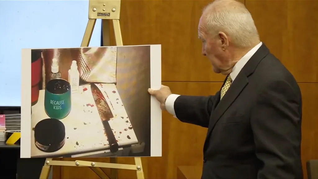 A man in a suit displays a crime scene photo showing a bloody knife, a cup that reads 