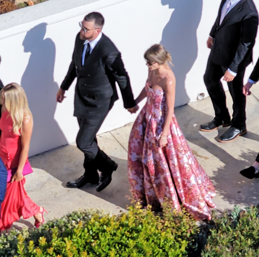 Taylor Swift in a pink floral dress holding hands with Travis Kelce in a suit.