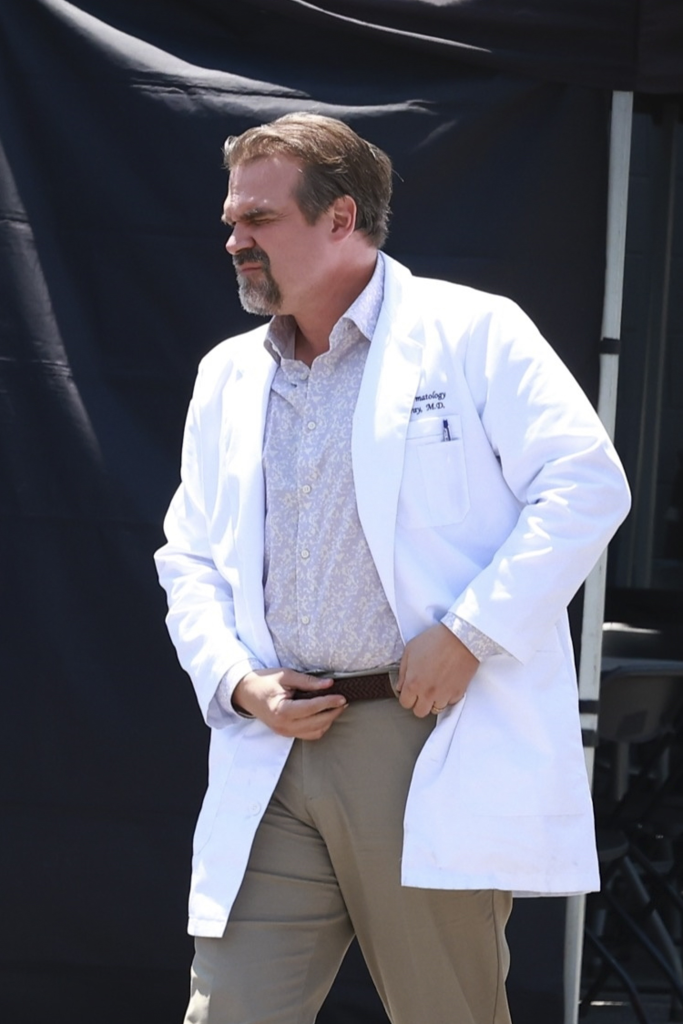 David Harbour as a doctor in a white lab coat and light-colored patterned shirt, adjusting his belt.