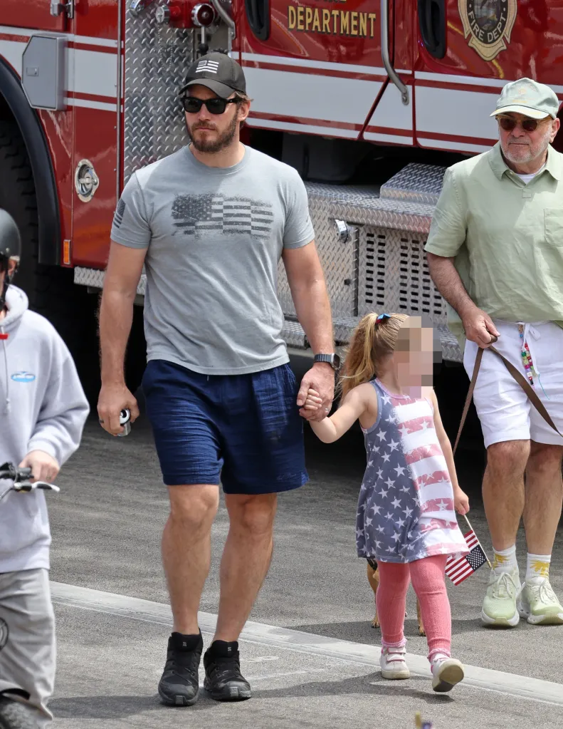 Chris Pratt holds a child's hand while standing in front of a fire engine at a 4th of July Parade.