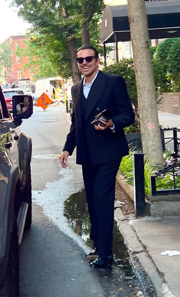 Bradley Cooper smiling as he stands on a wet street next to a dark vehicle.