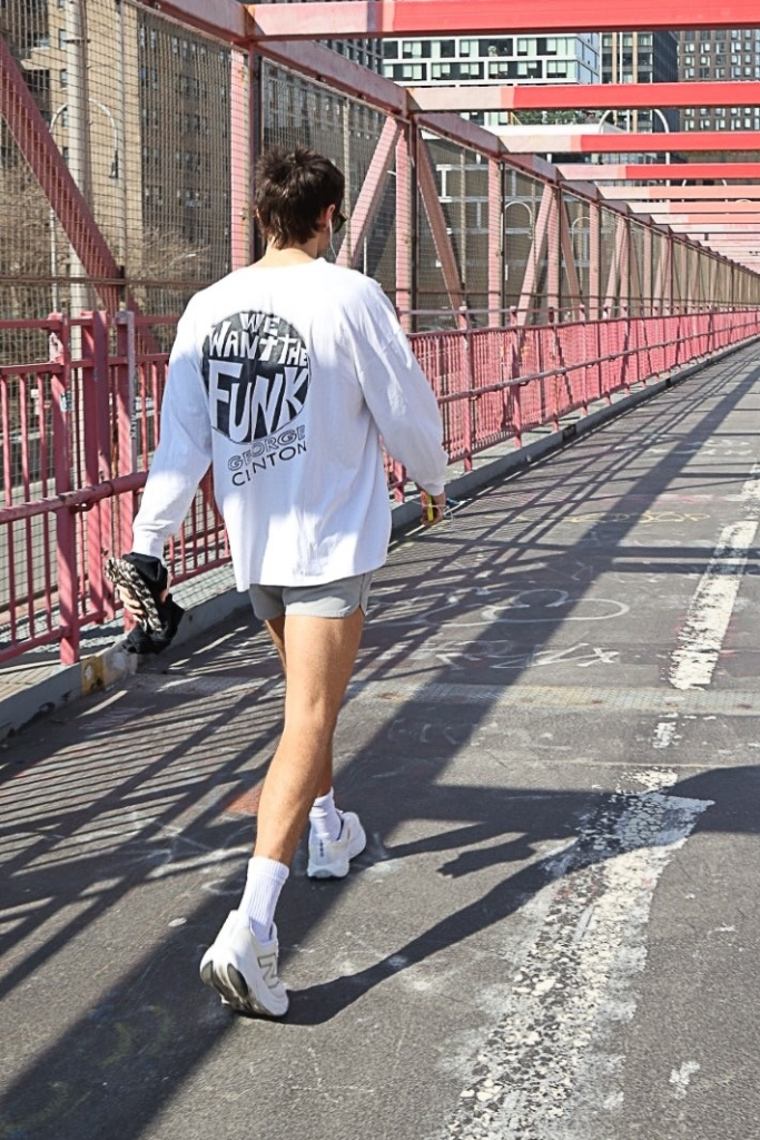 Harry Styles walking on a bridge in New York City wearing a white long-sleeved shirt, gray shorts, and white sneakers.