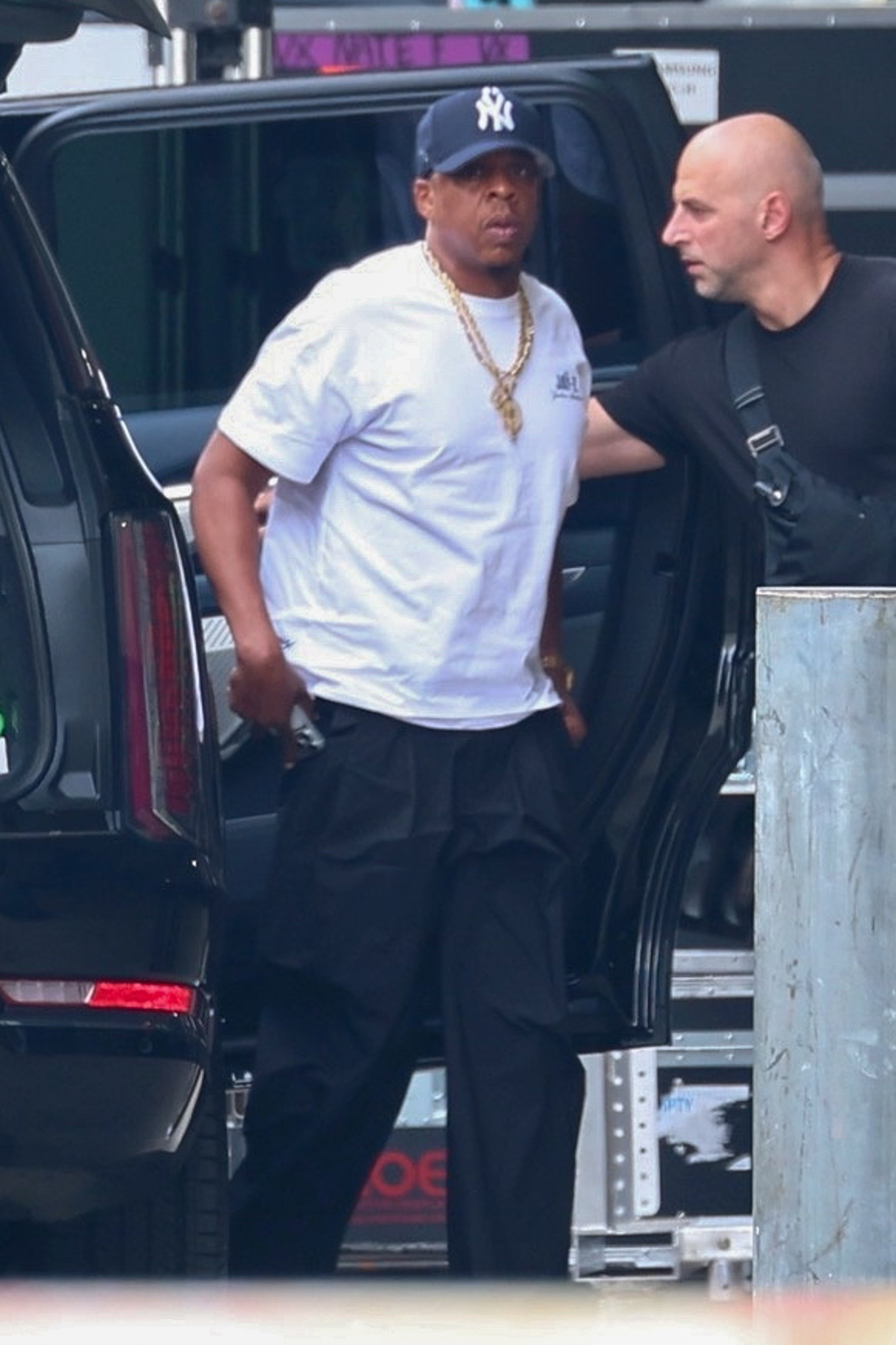 Jay-Z arriving at Yankee Stadium, wearing a New York Yankees cap, a white t-shirt, and gold chains, with a man in a black shirt beside him.