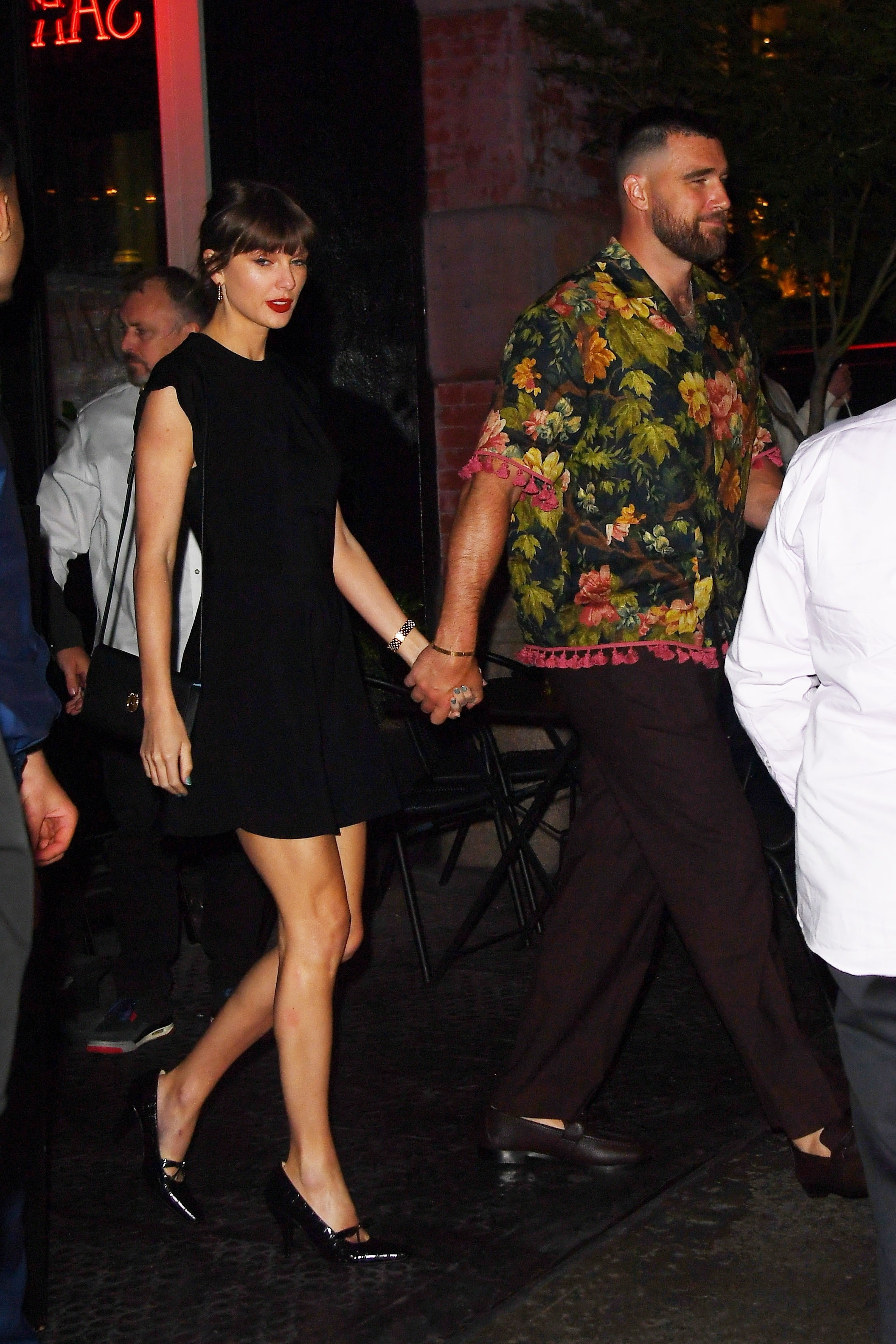 Taylor Swift in a black dress and Travis Kelce in a floral shirt, holding hands after dinner.