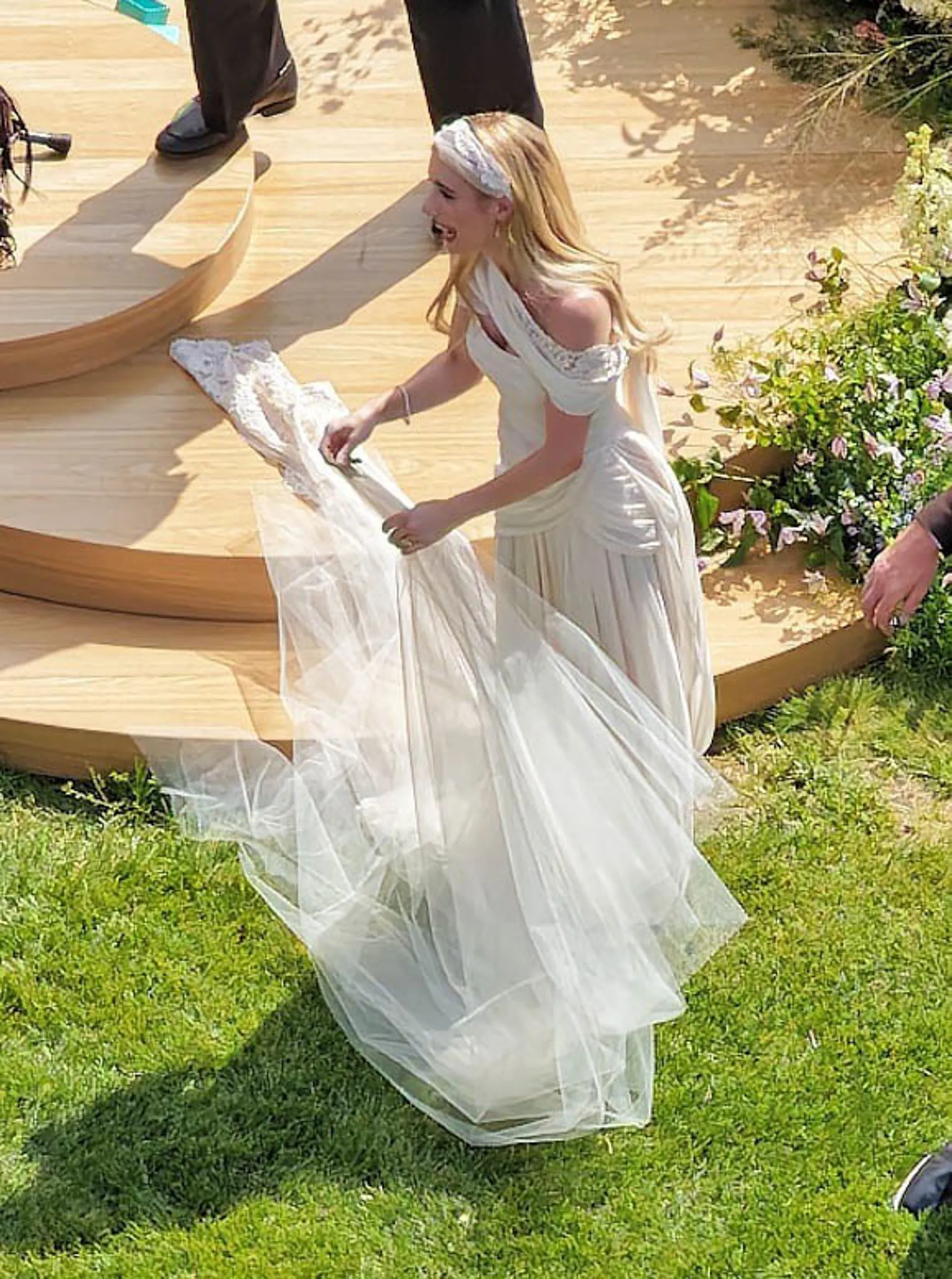 Emma Roberts in a white dress, veil, and headband, smiling while standing on a wooden platform next to green grass.