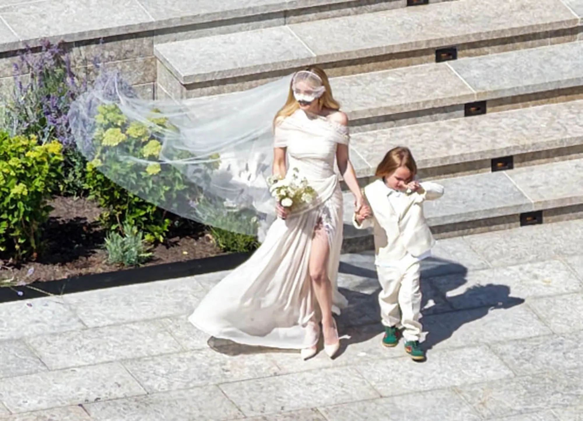 Emma Roberts in her white wedding gown and veil, holding hands with a young boy in a white suit, walking on a stone patio.