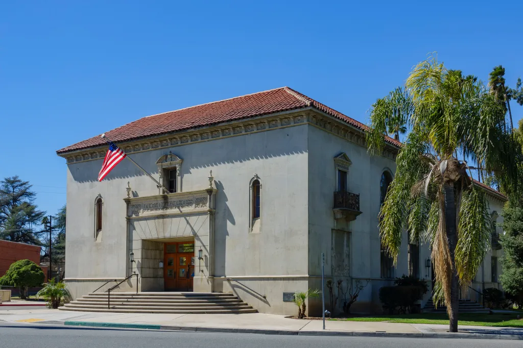 Exterior view of the Redlands Police Department Administrative Office, a light-colored building with a terracotta tiled roof, an American flag, and a palm tree in front.