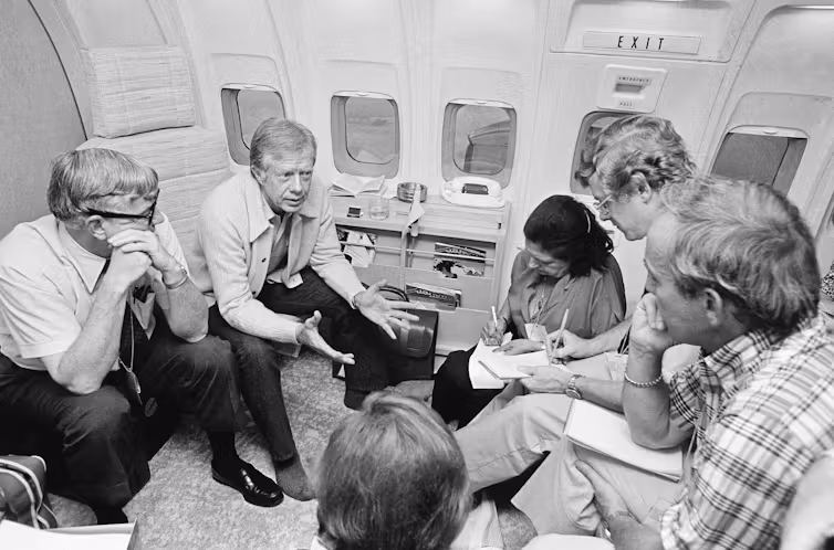 A group of people seated on an airplane listening to a man speaking and gesturing with his hands.