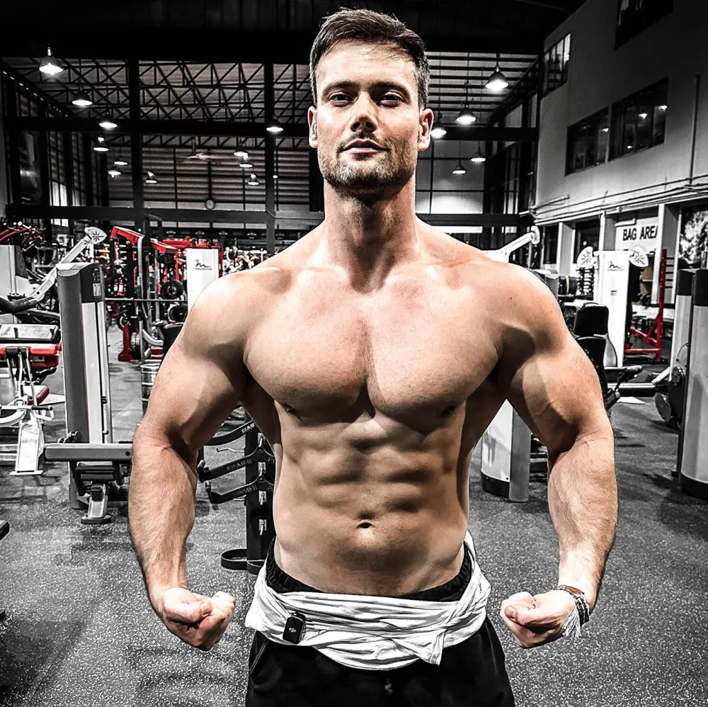 Connor Michael Murphy, a muscular man with a beard and dark hair, flexing in a gym.