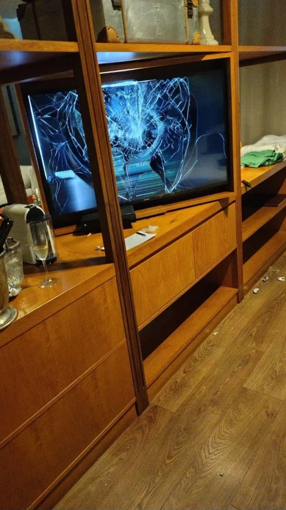 Hotel room with a shattered TV screen, an empty champagne glass, and items scattered on the floor, suggesting destruction, drugs, and alcohol.