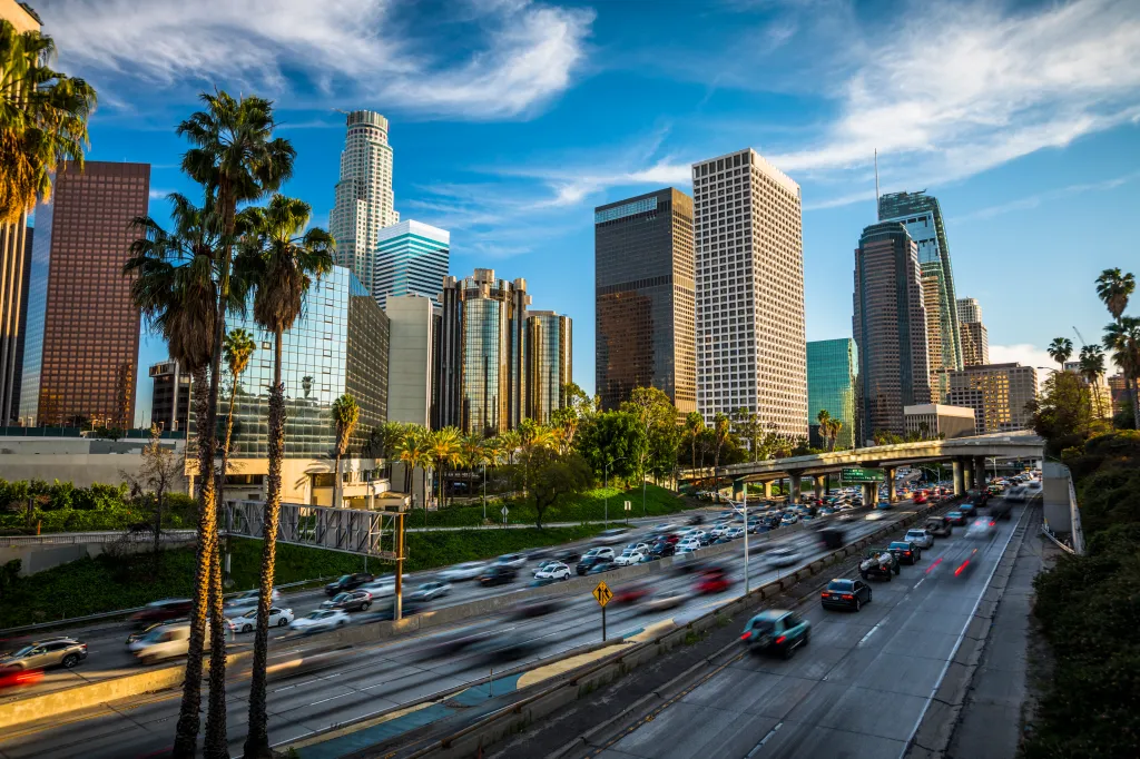 Daytime shot of a Los Angeles freeway during rush hour, with tall skyscrapers and palm trees in the background.