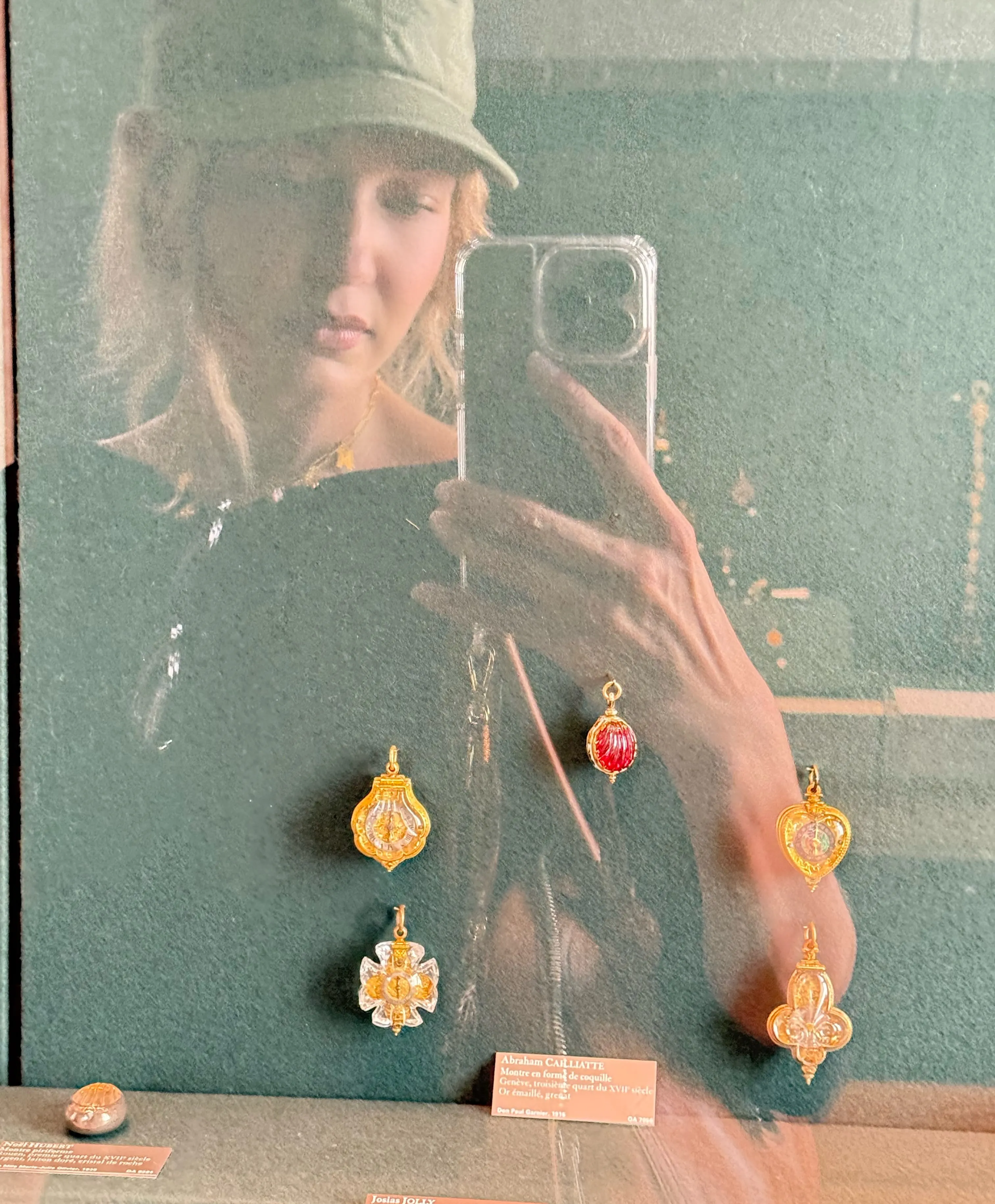 Gigi Hadid taking a selfie reflecting on a museum glass display case holding miniature decorative clocks.