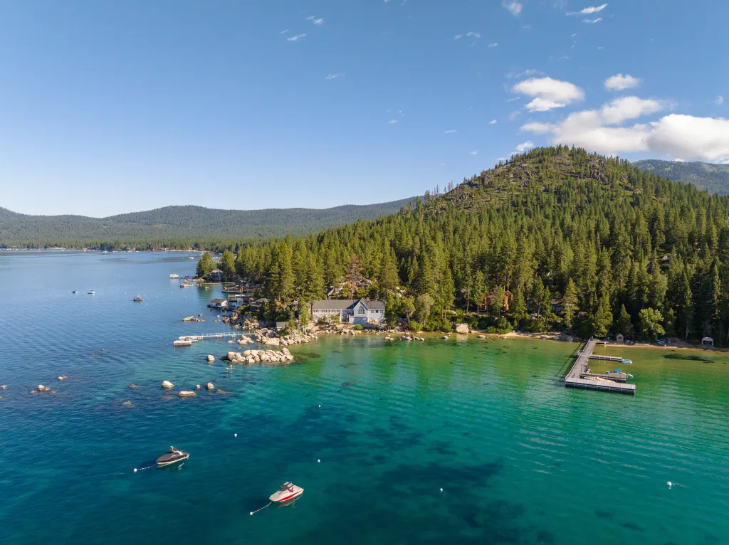 Aerial view of a house nestled among pine trees on the shore of Lake Tahoe, with clear blue-green water and a long wooden pier.