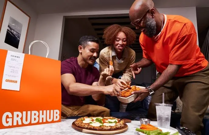 Three people sharing Grubhub food, including pizza and chicken wings, with a large orange Grubhub bag in the foreground.