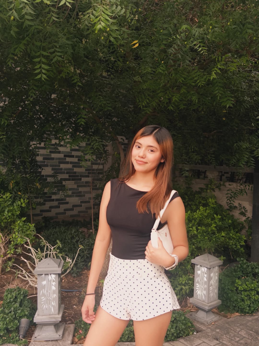 A young woman in a black top and polka-dot shorts stands outdoors with trees and bushes in the background.