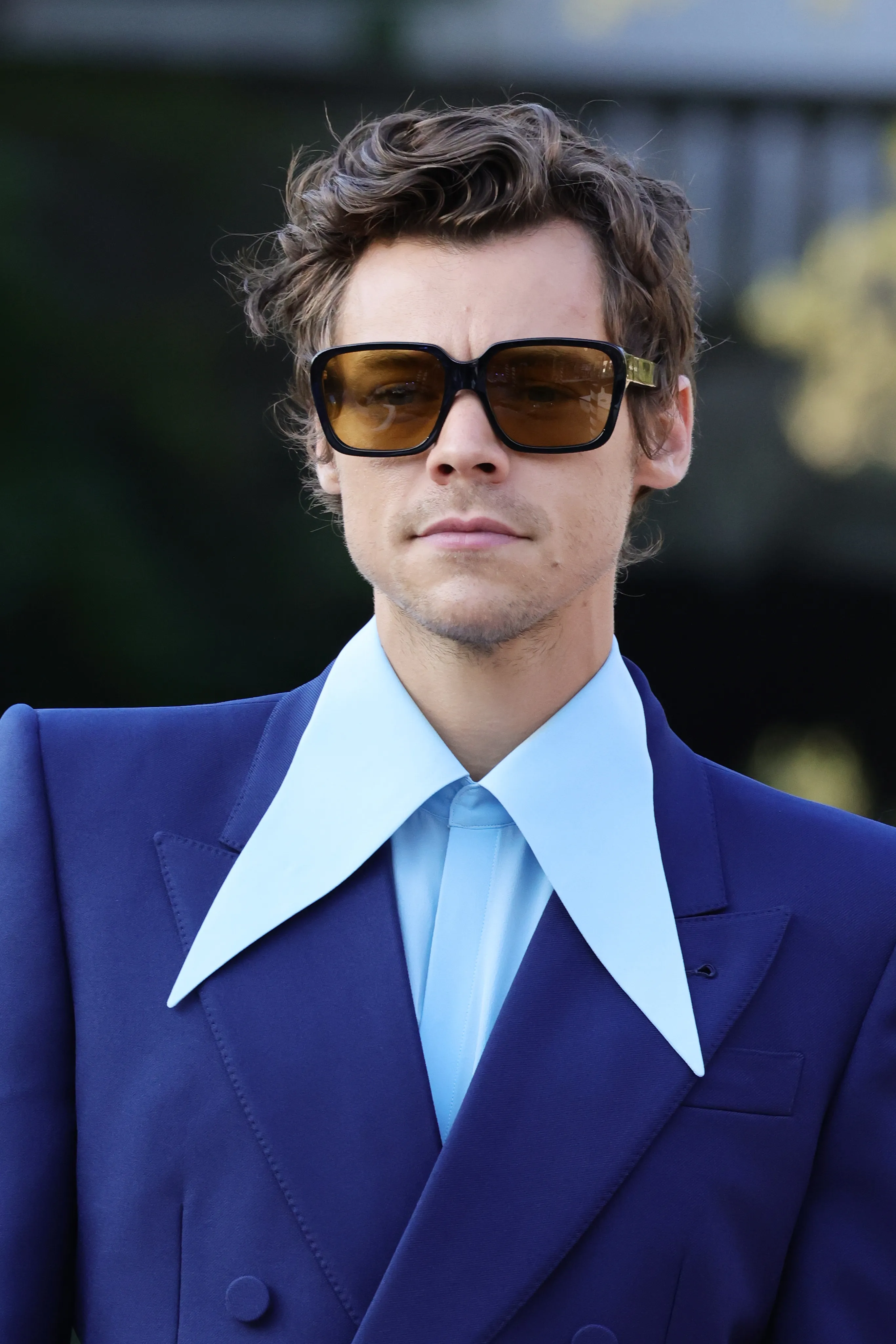 Harry Styles wearing a blue suit, light blue shirt with a wide collar, and sunglasses.