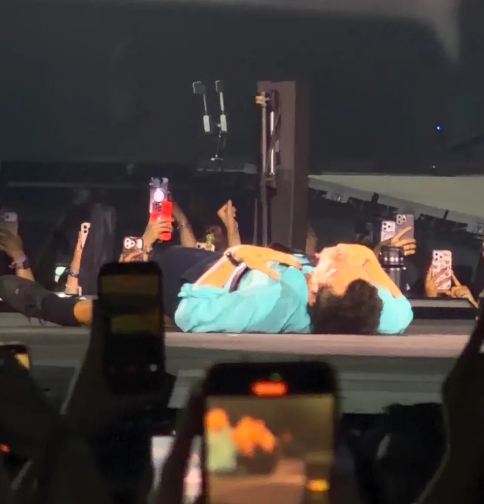 Harry Styles, in a blue shirt and black shorts, lying on stage with hands on his head, surrounded by fans holding up phones.