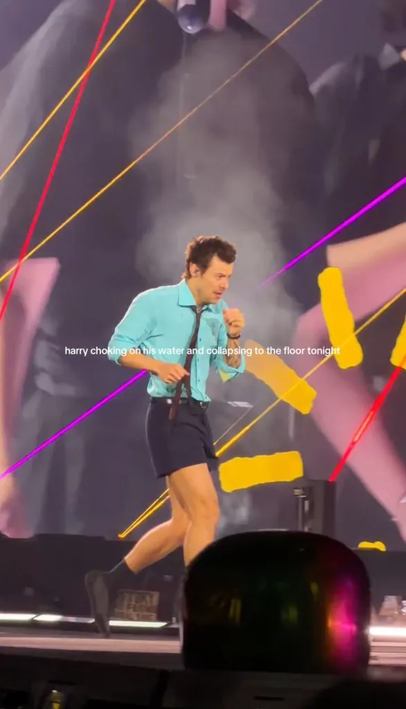 Harry Styles on stage, wearing a blue shirt and dark shorts, appearing to choke on water.