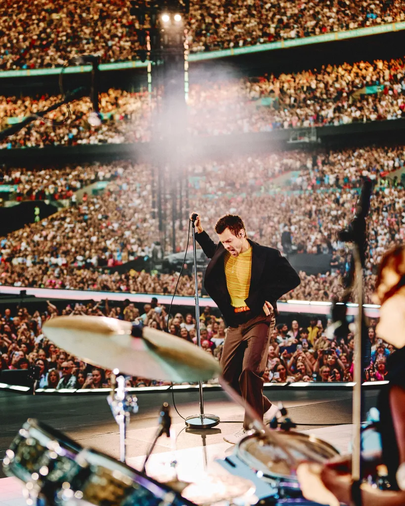 Harry Style in a black jacket, yellow top and brown pants, performing on stage at Wembley Stadium, holding a microphone with a large crowd in the background.