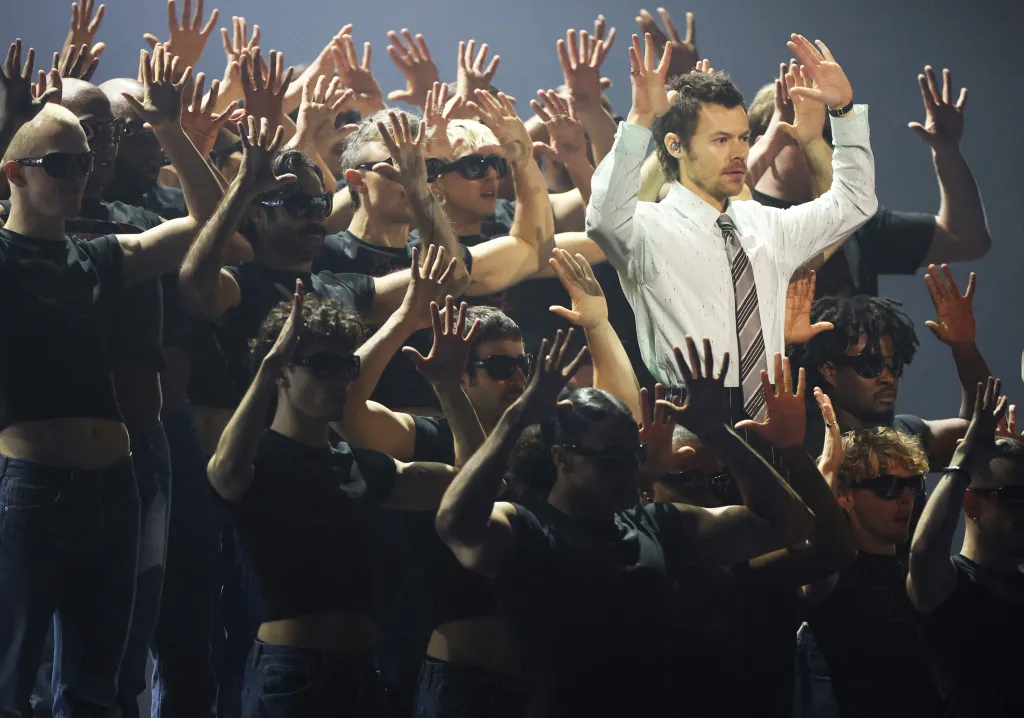 Harry Styles, in a white shirt with black and grey striped tie, performing on stage at the BRIT Awards surrounded by dancers with their hands raised.
